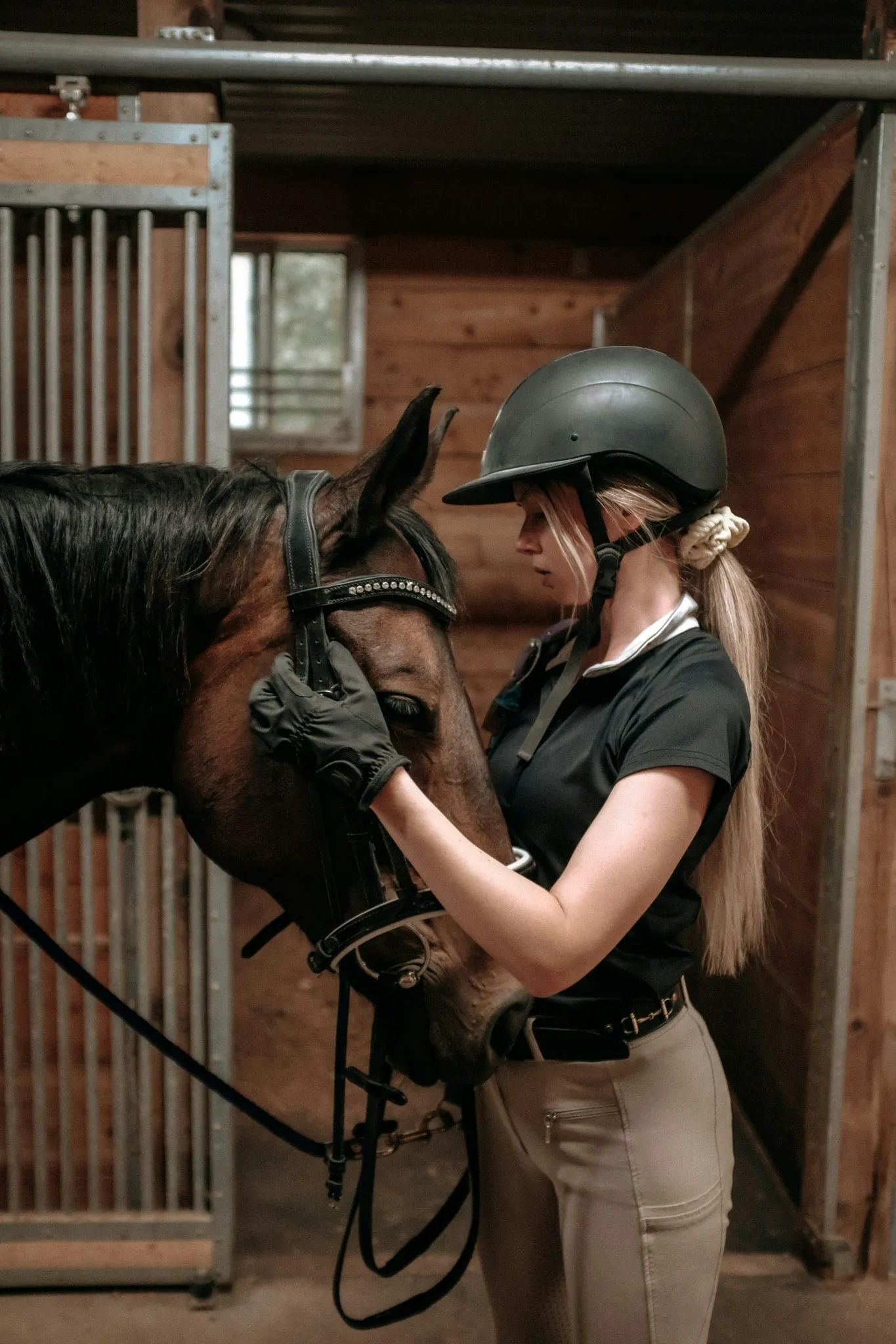 Helmeted rider standing beside a bridled bay horse in a stable